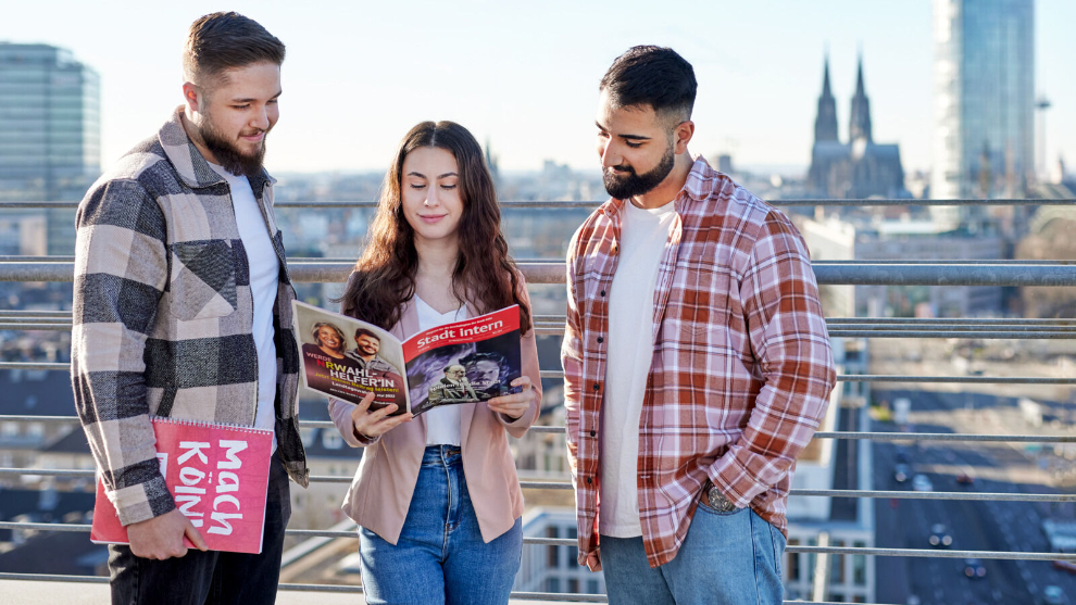 Drei Azubis stehen auf einer Dachterrasse und blättern in der Stadt Intern, im Hintergrund ist der Dom zu sehen.