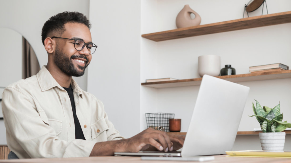 Junger Mann mit Brille sitzt lächelnd mit Laptop am Tisch