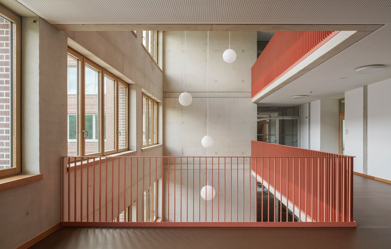 Blick in das helle, offene Treppenhaus mit rötlichem Boden, rotem Geländer und Hängelampen in Kugelform