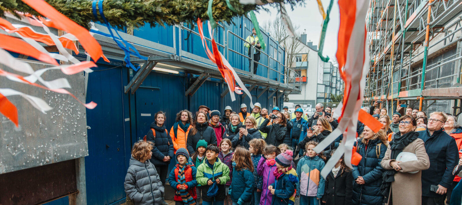 Schüler*innen der Grundschule mit Feiergesellschaft der am Bau Beteiligten, unter anderem Architekt Christian Fensterer und technische Betriebsleiterin der Gebäudewirtschaft Petra Rinnenburger, beim Hochziehen des Richtkranzes