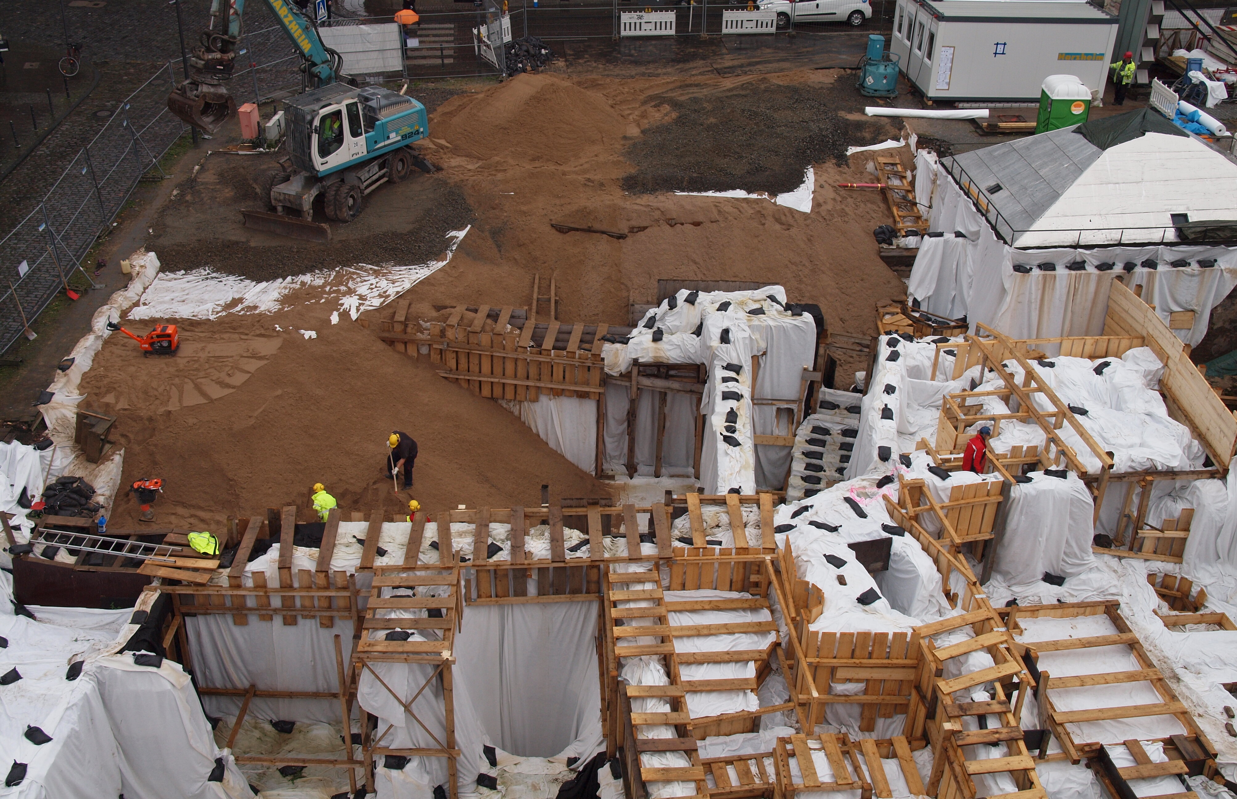 Ein Schaufelbagger und Männer mit Schaufeln bringen den Sand in die Archäologische Zone ein.