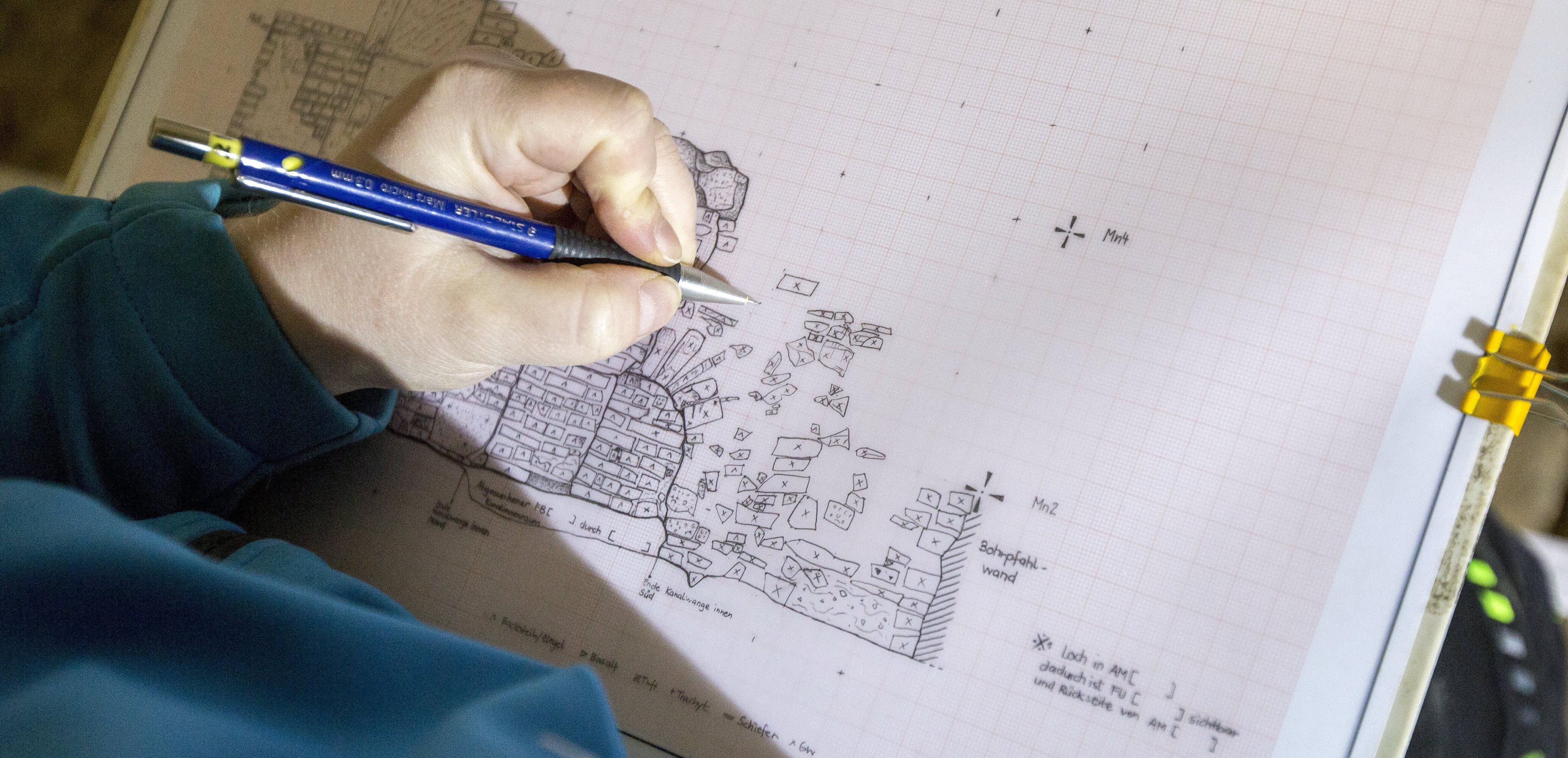 Nahaufnahme der linken Hand eines Mitarbeitenden der Archäologie, der mit einem sehr feinen Bleistift eine filigrane Zeichnung vieler kleinen Felder anzeichnet, um zu dokumentieren, wo welcher Fund gesichert wurde.