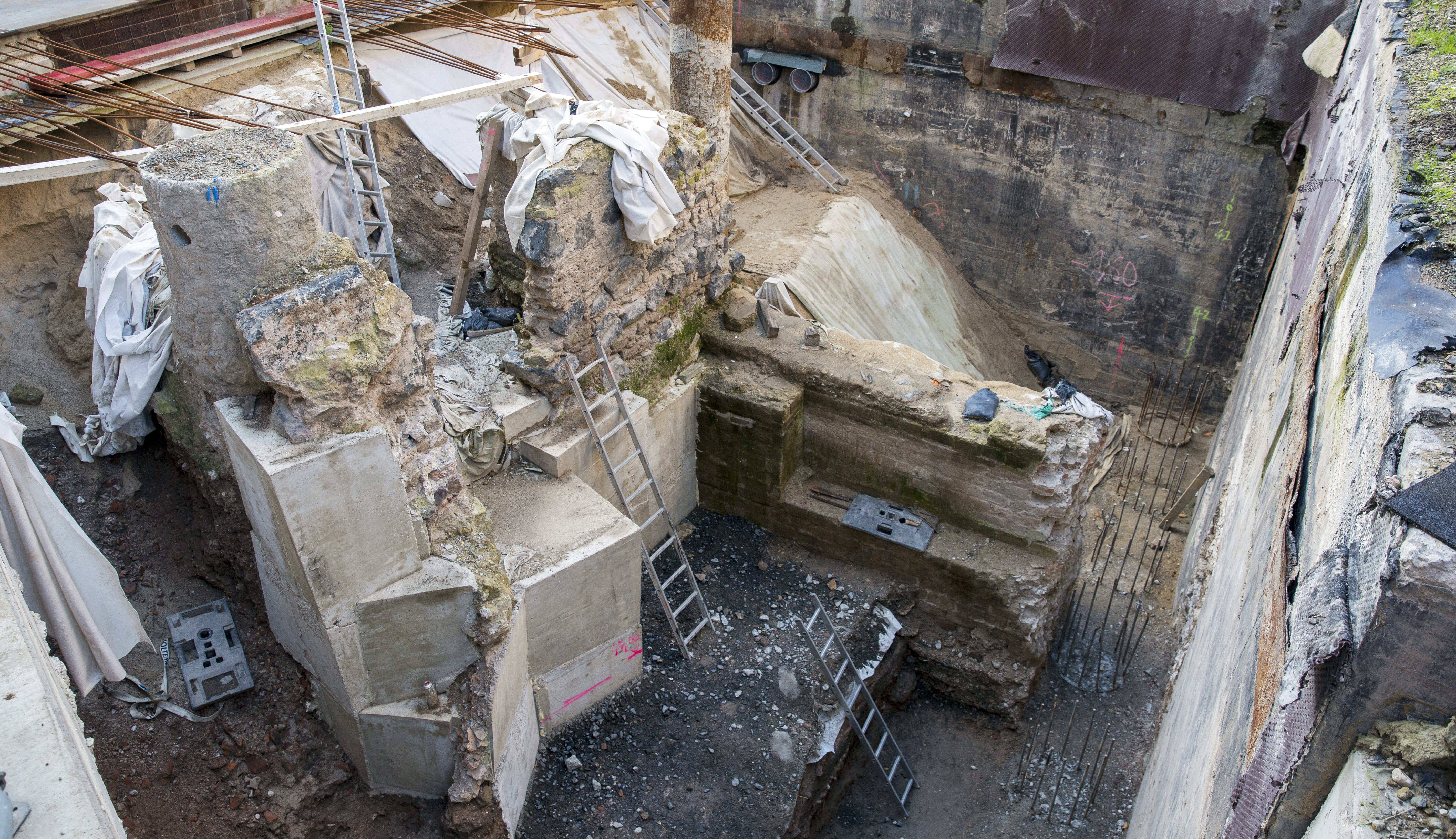 Metertief unter den ergrabenen historischem Mauerwerk wurden blöckeweise Unterfangungen aus Beton eingebracht, um die Statik zu sichern. Mehrere Leitern führen über die mehrere Ebenen bis ganz nach unten.