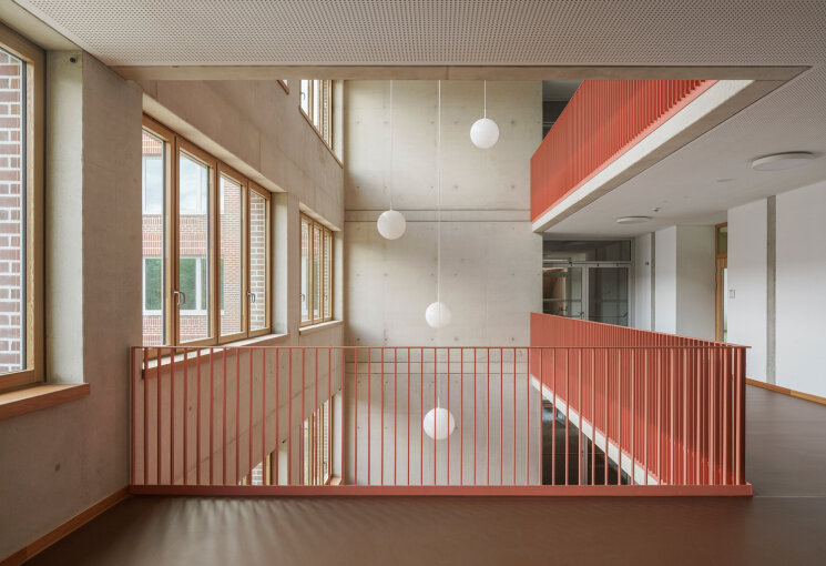 Blick in das helle, offene Treppenhaus mit rötlichem Boden, rotem Geländer und Hängelampen in Kugelform, © Sebastian Schels