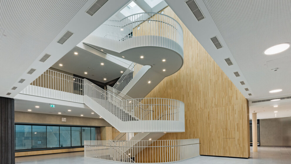 Durch ein Atrium fällt sehr viel Tageslicht in ein Foyer mit offenem Treppenhaus, das an den Wänden mit hellem Holz verkleidet wurde. Dahinter eine Sitznische mit Fensterreihe, die Blicke in die darunter liegende Sporthalle ermöglicht.