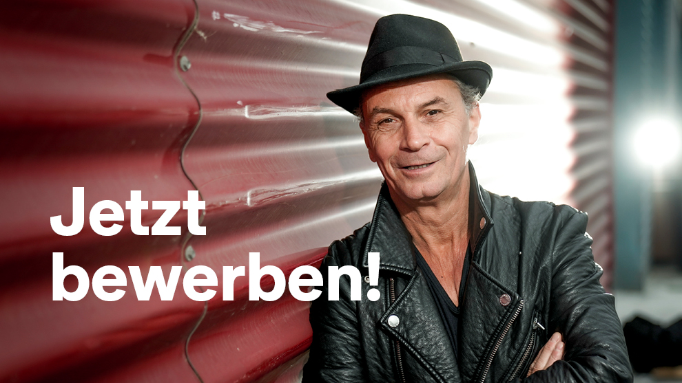 Das Foto zeigt den Ehrenamtspaten 2026 Peter Brings in schwarzer Lederjacke und mit schwarzem Hut, der sich an eine Mauer anlehnt. Im Hintergrund ist ein Scheinwerfer zu sehen, der Peter Brings von hinten anleuchtet. Vor Ihm befindet sich ein weißer Schriftzug: Jetzt bewerben!