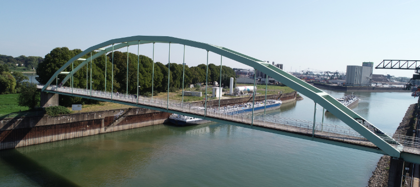 Zu sehen ist die Niehler Hafenbrücke mit Industrieanlagen im Hintergrund sowie zwei Tankern auf dem Rhein.