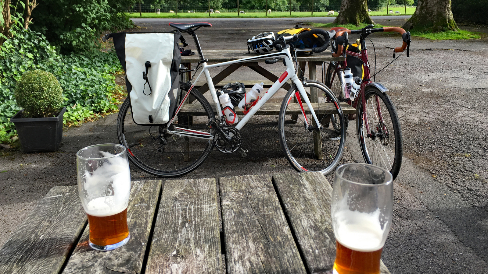 Auf einem Holztisch stehen zwei angetrunkene Gläser Bier. Im Hintergrund lehnen zwei Fahrräder an einer Bank., &copy Harry Green – stock.adobe.com