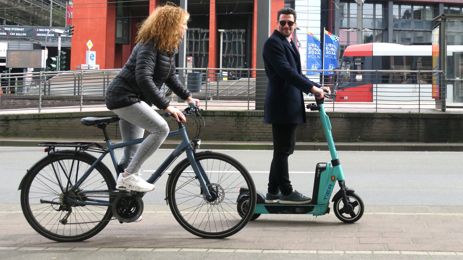 Eine Radfahrende wird auf einem Radweg von einem lächelnden E-Roller-Fahrer überholt., &copy Stadt Köln