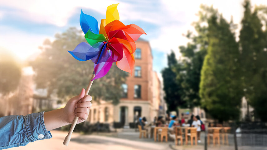Windrädchen in Kinderhand. Im Hintergrund sieht man die Fassade eines Hauses mit Menschen auf Stühlen davor.